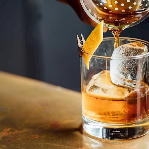 A glass of amber whiskey with ice, a citrus twist garnish, and a pour being added from a decanter, on a wooden bar.