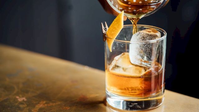 A glass of amber whiskey with ice, a citrus twist garnish, and a pour being added from a decanter, on a wooden bar.