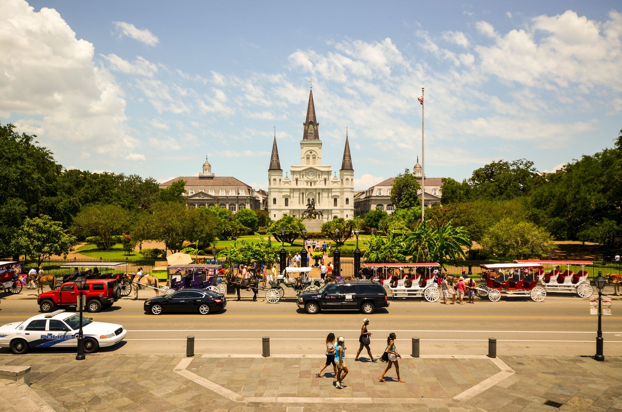 Medieval-style castle-like building with spires in the background, a busy plaza, people walking, and cars on a street in front.