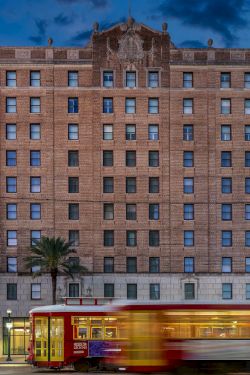 A city street at dusk with a tall brick building, a blurred red trolley in motion, palm trees, and surrounding modern structures.