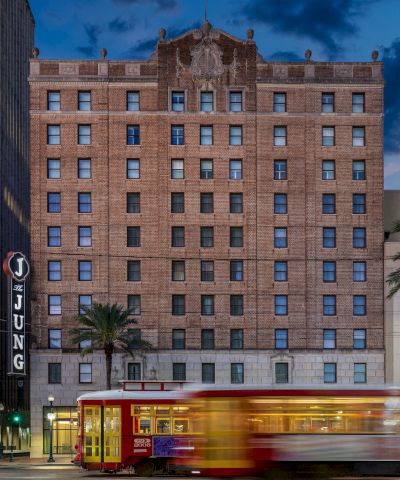 A city street at dusk with a tall brick building, a blurred red trolley in motion, palm trees, and surrounding modern structures.