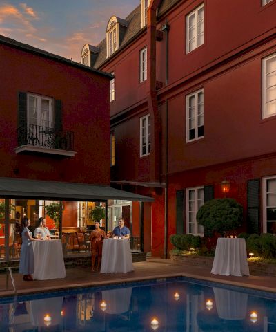 A courtyard hotel bar by a pool at dusk, with guests at high-top tables under a shaded canopy, red brick buildings surrounding.