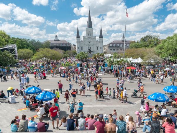 A large crowd gathers in a town square with a grand white church behind, blue umbrellas, stalls, and people strolling, enjoying a sunny day.