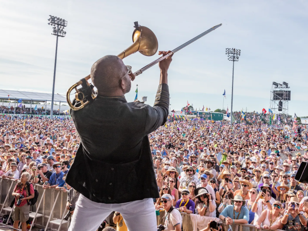A musician on stage blasting a brass trumpet into a huge outdoor crowd, crowd surfers and cheering fans fill the festival grounds.