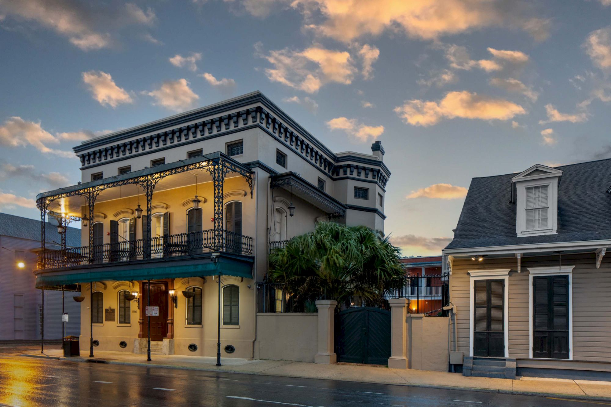A vintage two-story building with a wraparound balcony, lights on, next to a small house, on a wet street at dusk.