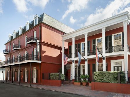 Colorful street with a red-brick building and white-columned portico; flags line the entrance, small potted shrubs, sunny sky.