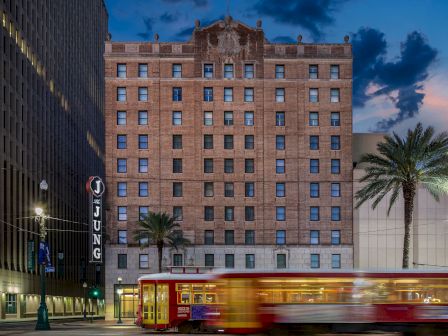 A street scene at dusk with a tall brick building, a red trolley bus in motion, palm trees, and illuminated storefronts along a city block.