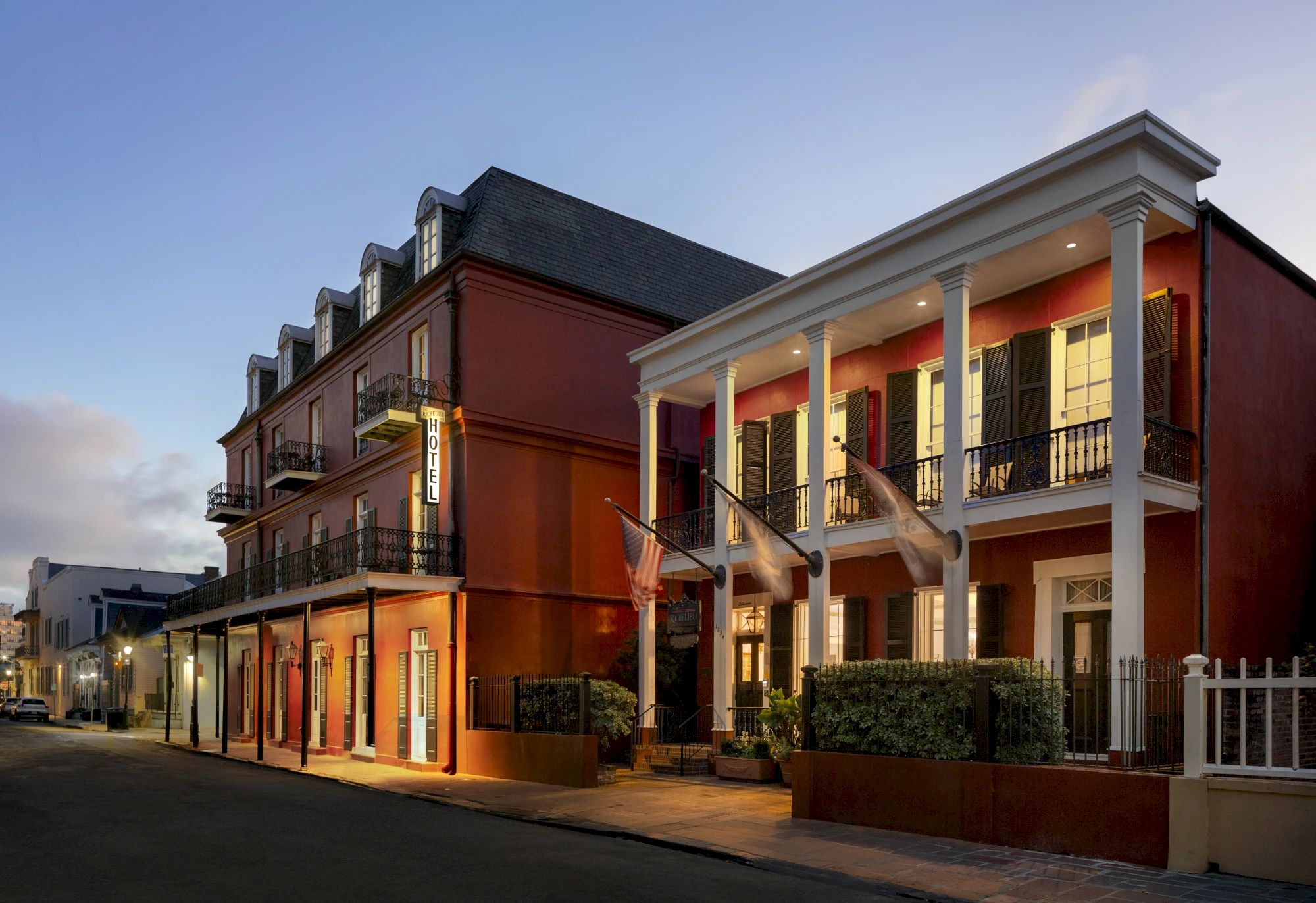 A row of charming brick townhouses with balconies and a covered porch, lit warmly as dusk settles on a quiet street.