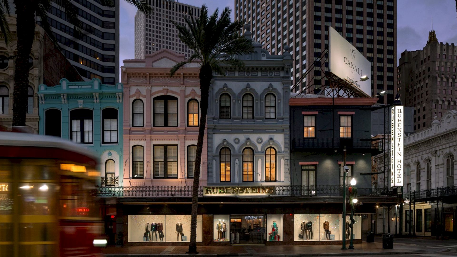 A row of colorful old-fashioned storefronts along a city street at dusk, with a red trolley in motion and palm trees framing the scene.