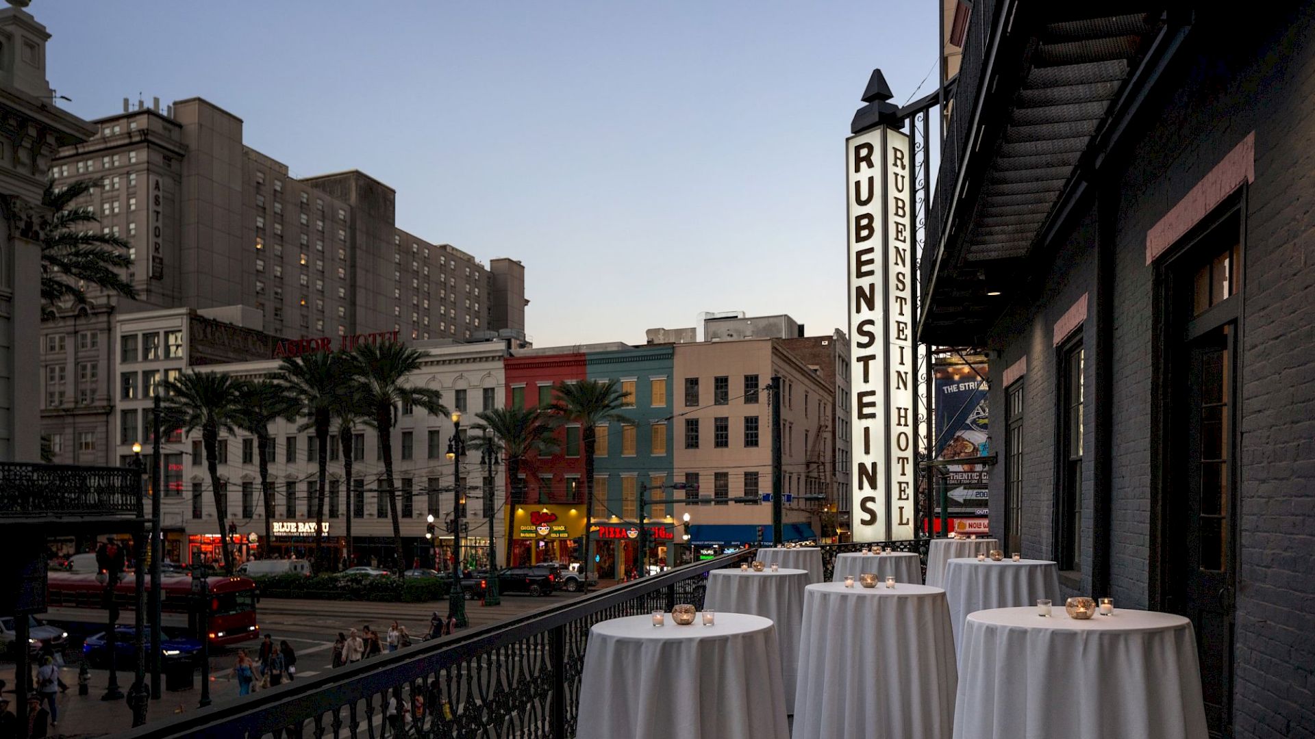 A downtown street scene with tall buildings, palm trees, and outdoor dining tables beside a vertical &ldquo;Restaurant&rdquo; sign.