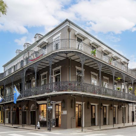 A classic corner building with two stories and wraparound wrought-iron balconies, storefronts on the ground level, and a bright blue sky above.