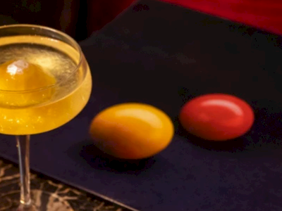 A cocktail glass with a yellow drink and ice, beside three colorful balls (orange and red), on a dark mat.