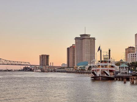 A riverfront skyline with a tall skyscraper, a bridge, and docked boats under a pastel sunset sky.
