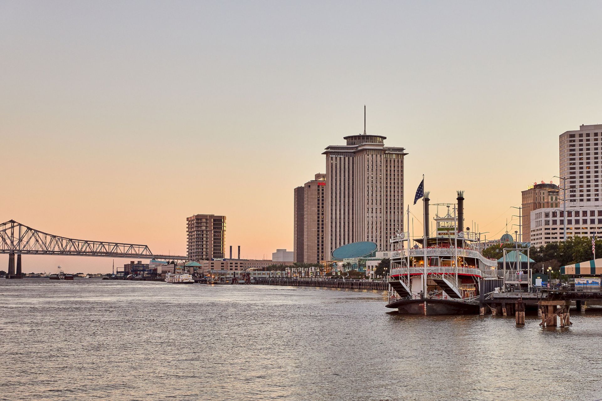 A riverside skyline with a tall office building, an American flag on a docked boat, and a long bridge at sunset.