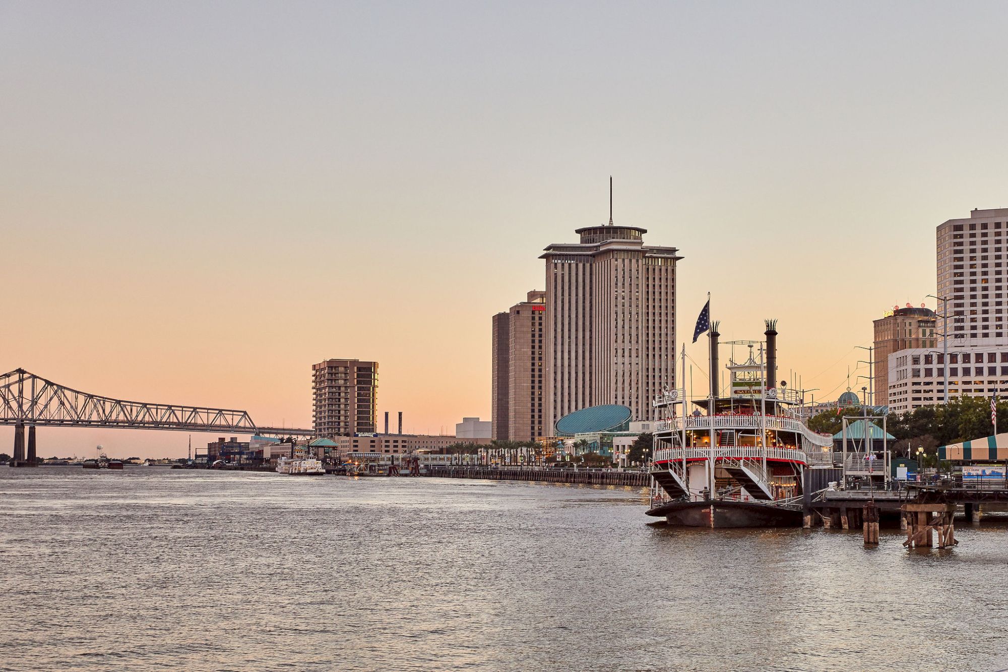 A riverside skyline with a tall office building, an American flag on a docked boat, and a long bridge at sunset.