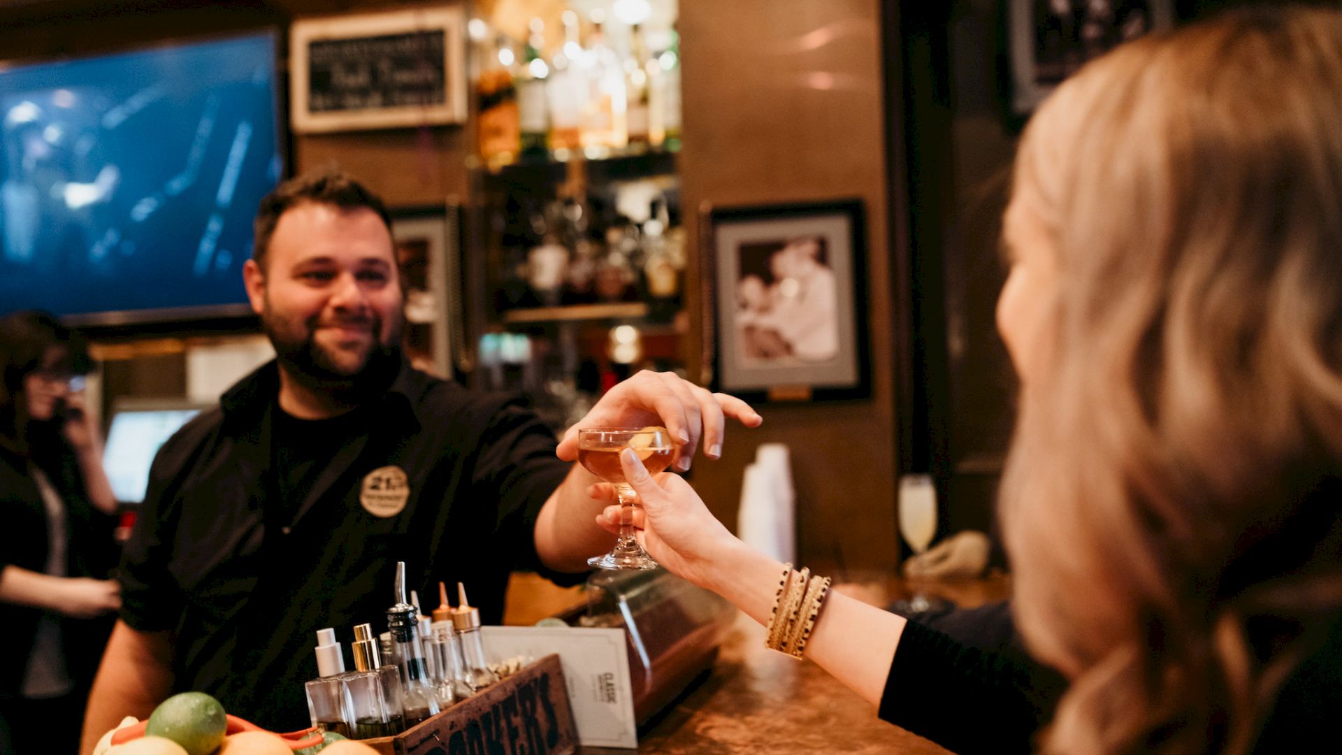 A bartender pours a drink into a guest&rsquo;s glass at a cozy bar, with bottles and taps lining the counter behind him.