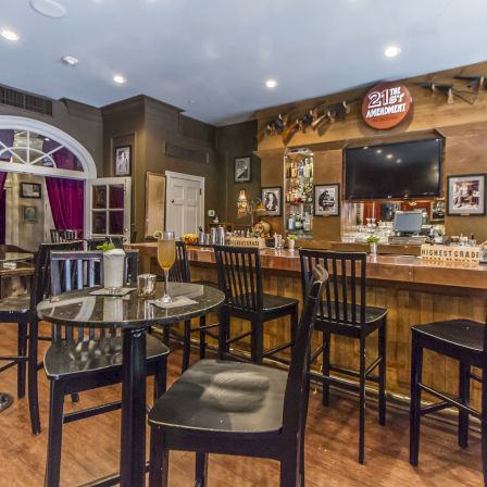 A cozy cafe/bar with wooden floors, high chairs around small glass tables, a well-lit bar area, and vintage photos on the walls. End.