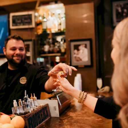 A bartender hands a customer a toast-ready cocktail at a warmly lit bar with drinks on the counter.