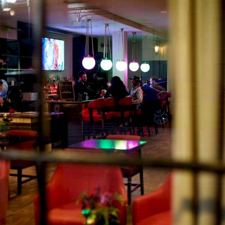 A cozy cafe interior with warm lights, red chairs, a few patrons, and a TV screen in the background.