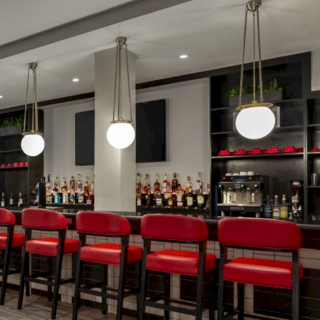 A modern bar with red cushioned stools, pendant lights, and a well-stocked back bar featuring bottles and glassware, ready for patrons.