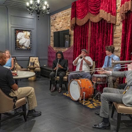 A small jazz band performs for an audience in a cozy room, with trombone, trumpet, clarinet, and drums under red curtains.