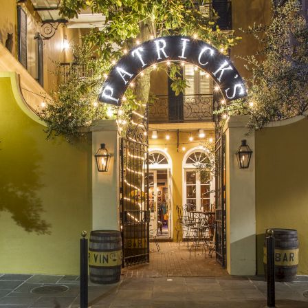 A warmly lit entrance to a quaint bar or restaurant named &ldquo;PATRICKS,&rdquo; with an arched sign, stone pathway, and hanging lanterns at night.