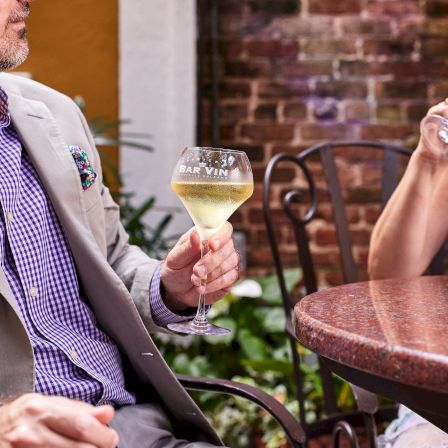 A stylish couple enjoying wine at an outdoor table; she sips white wine while he leans back in a beige blazer.
