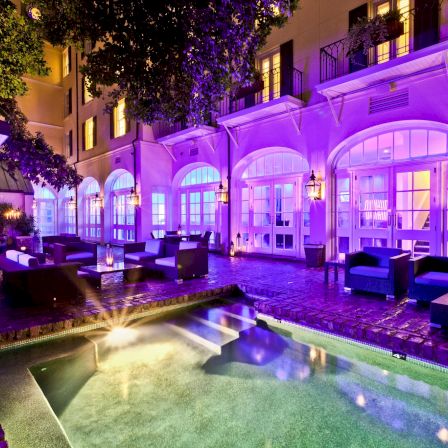 A luxurious outdoor hotel courtyard at night with a lit purple fa&ccedil;ade, comfortable seating, and a small pool reflecting colorful lights.