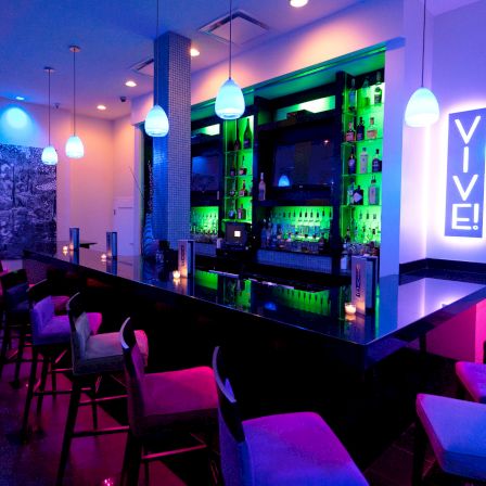 A modern bar with neon blue and purple lighting, a glossy black counter, stools, glowing shelves, and a sign that reads &ldquo;VIVE!&rdquo;