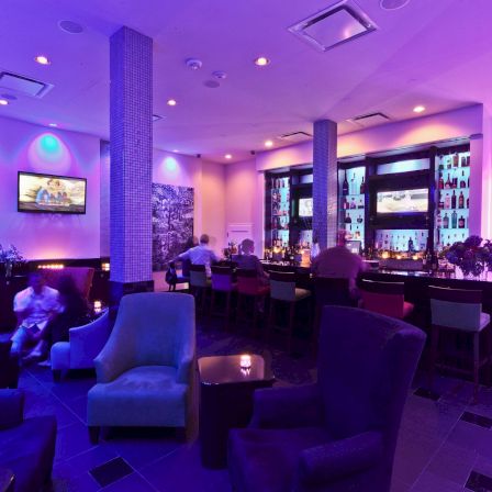 A chic, dimly lit lounge/bar with blue and purple lighting, plush seating, a long bar, and a cozy seating area for conversation.