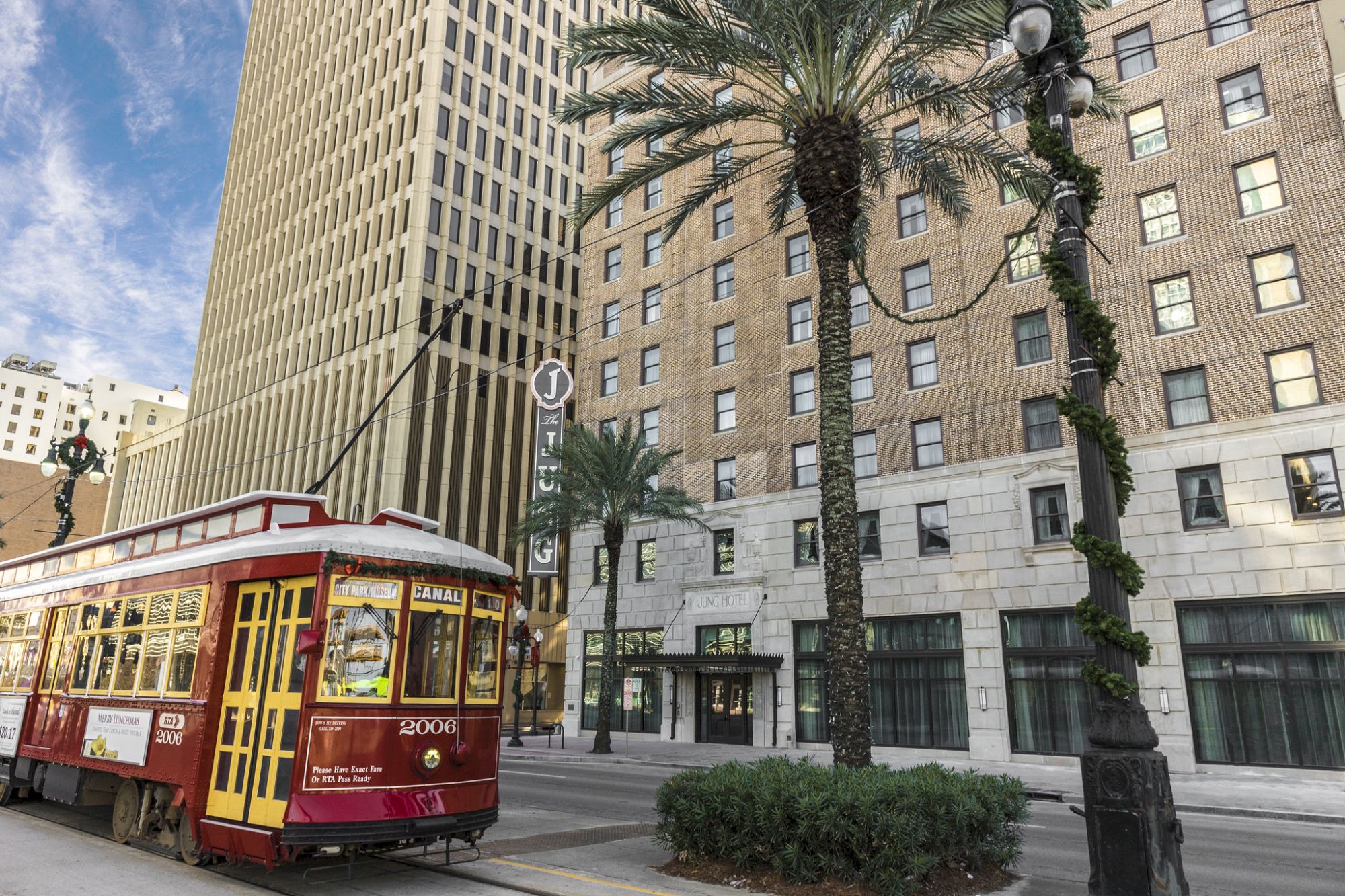 A red streetcar and a brick hotel building with palms and streetlights line a sunny city street.