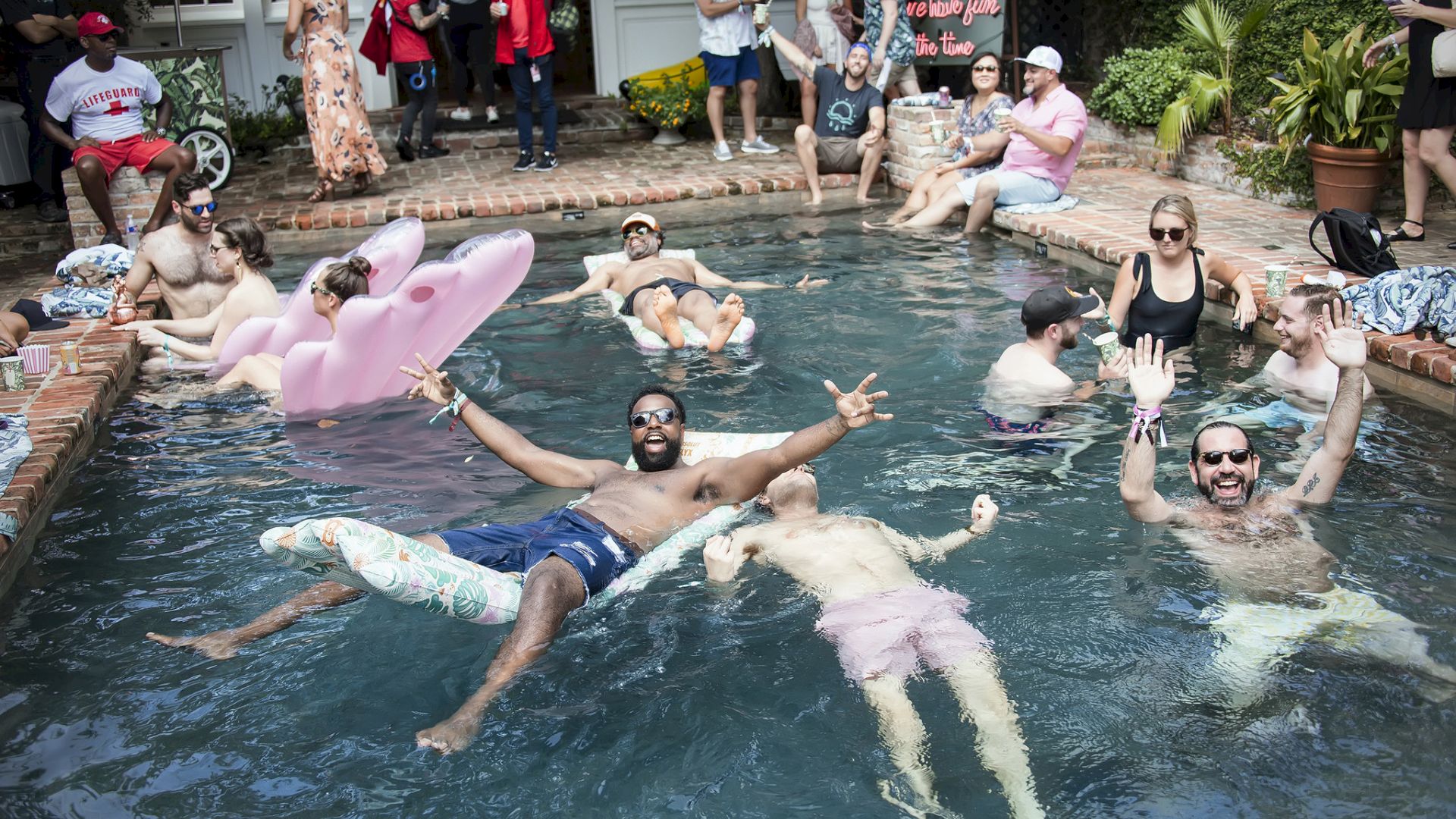 People swimming and lounging in a pool, some with inflatable toys, others posing and chatting on the edge, a casual backyard party vibe.