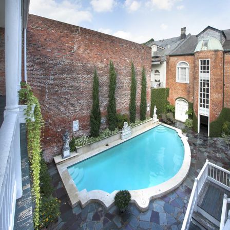 A small, elegant backyard with a curved turquoise pool, bordered by a tall brick wall, ivy, and slender trees, adjacent to brick buildings and a stairwell.