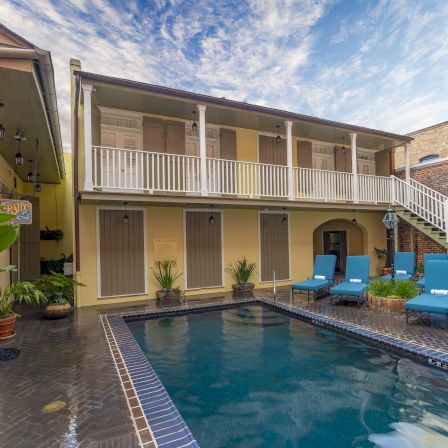 A cozy two-story courtyard with a rectangular pool, tropical plants, and blue lounge chairs under a sunny, partly cloudy sky.