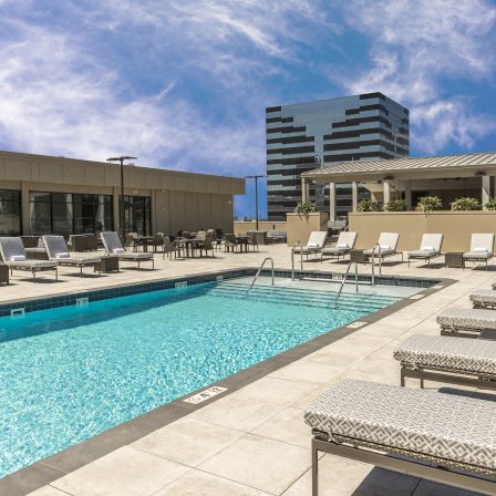 A rooftop hotel pool with crystal blue water, surrounding lounge chairs, and modern buildings in the background. The sky is clear and sunny.