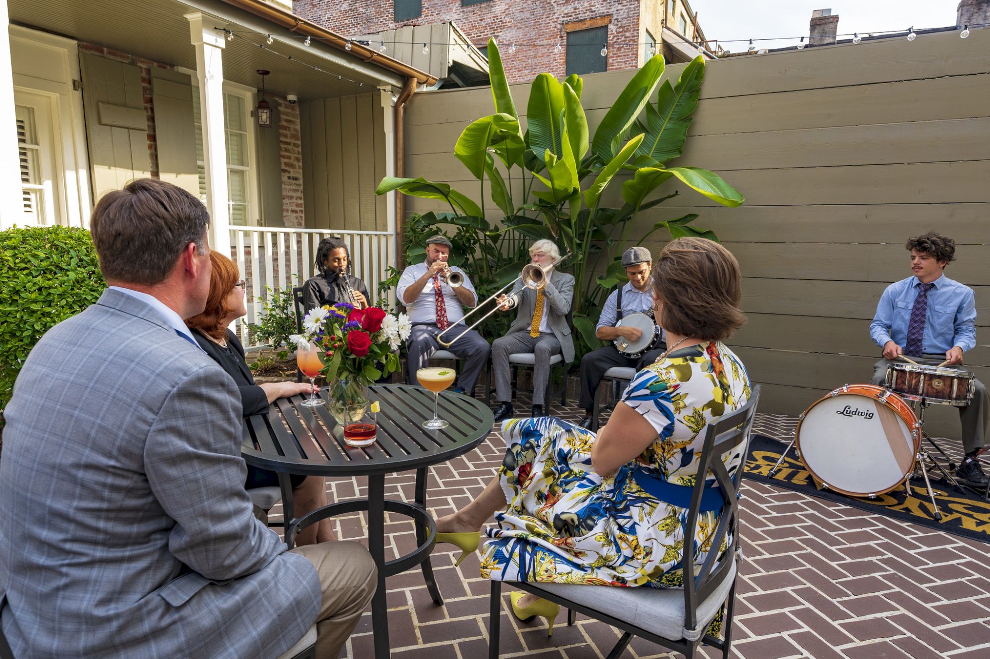 An outdoor patio party features musicians playing horns, banjo, and drums while guests enjoy drinks and flowers.