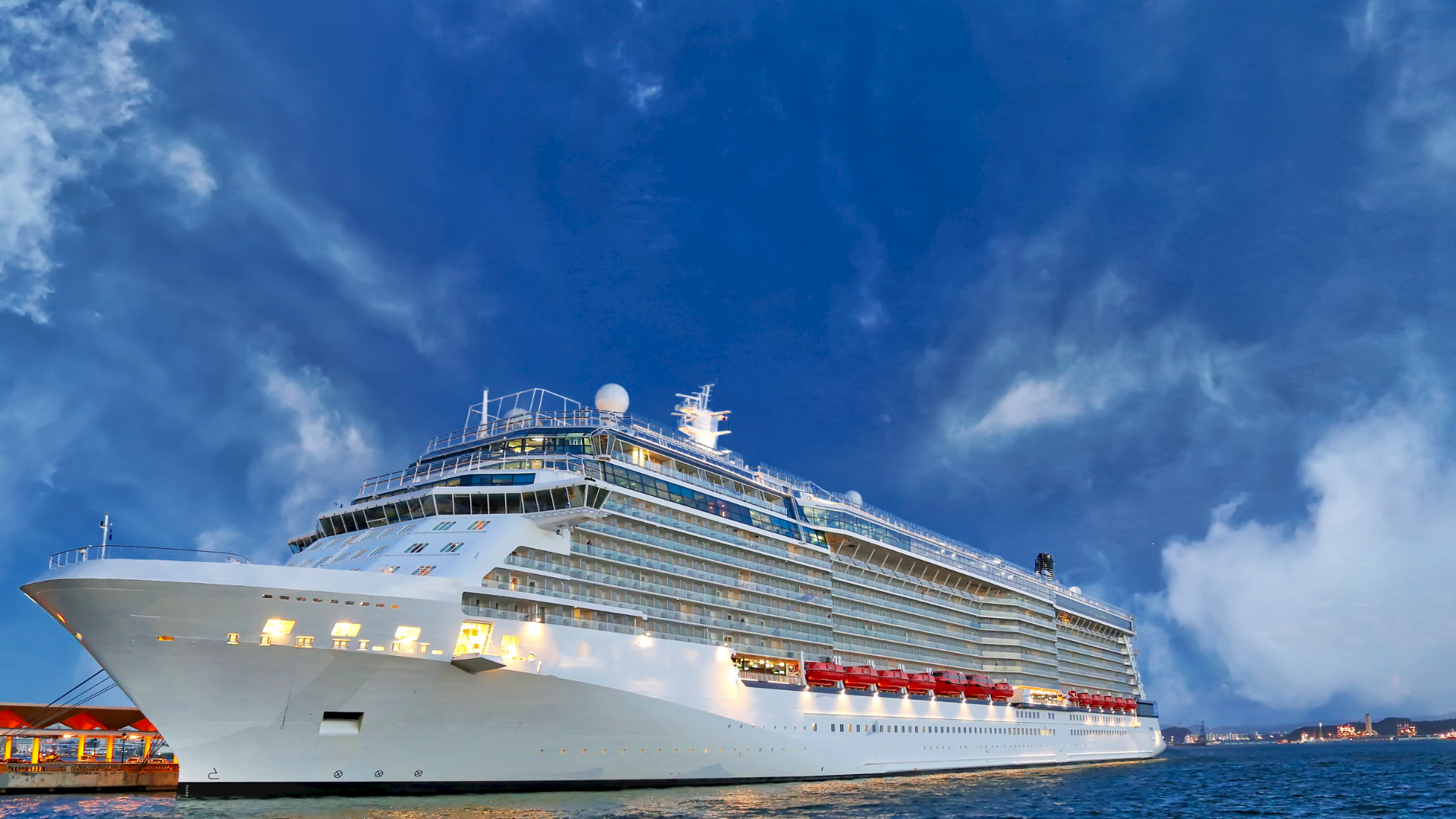 A large cruise ship sails at dusk under a bright blue sky with scattered clouds.