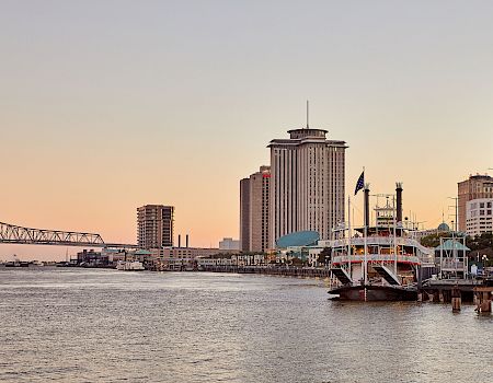 Sunset river skyline: tall tower, moored riverboat with flags, and a distant steel bridge over the water.