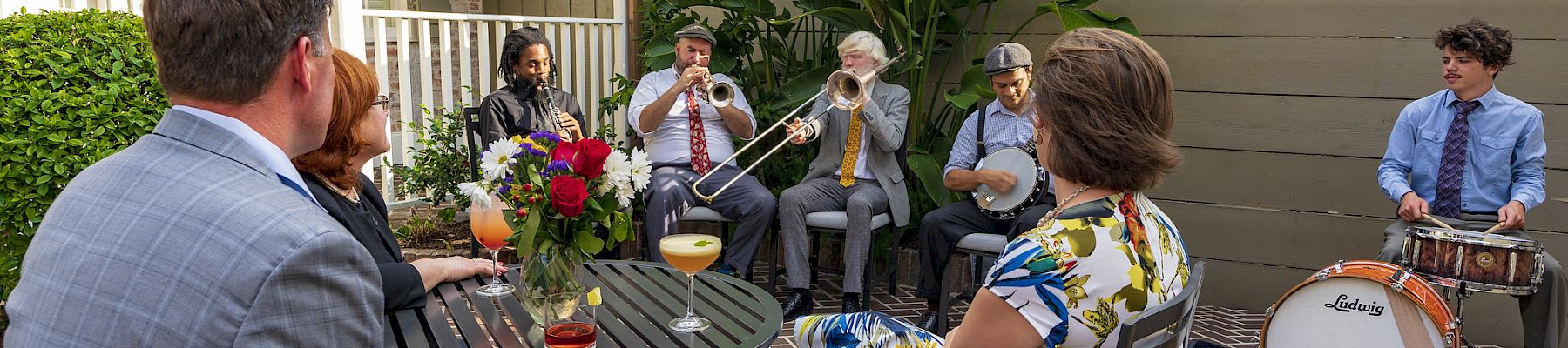 An outdoor patio party where a small band plays brass and drums for guests seated around a table with drinks and flowers.