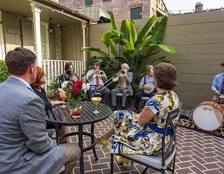 An outdoor patio party where a small band plays brass and drums for guests seated around a table with drinks and flowers.
