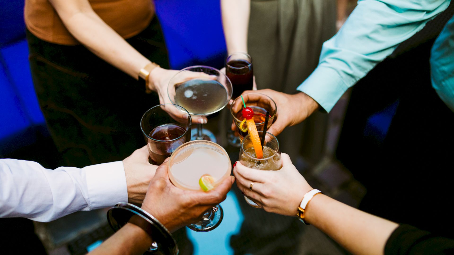 People clink glasses of drinks at a gathering, raising cocktails with citrus and cherries.