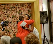 At a wedding-style event, a woman in orange laughs while hugging someone near a floral backdrop and a microphone speaker.