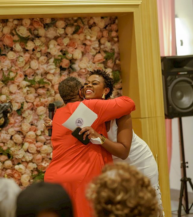 At a wedding-style event, a woman in orange laughs while hugging someone near a floral backdrop and a microphone speaker.