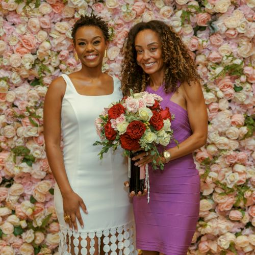 Two women in elegant dresses stand smiling against a wall of roses, holding a bouquet for a special occasion.
