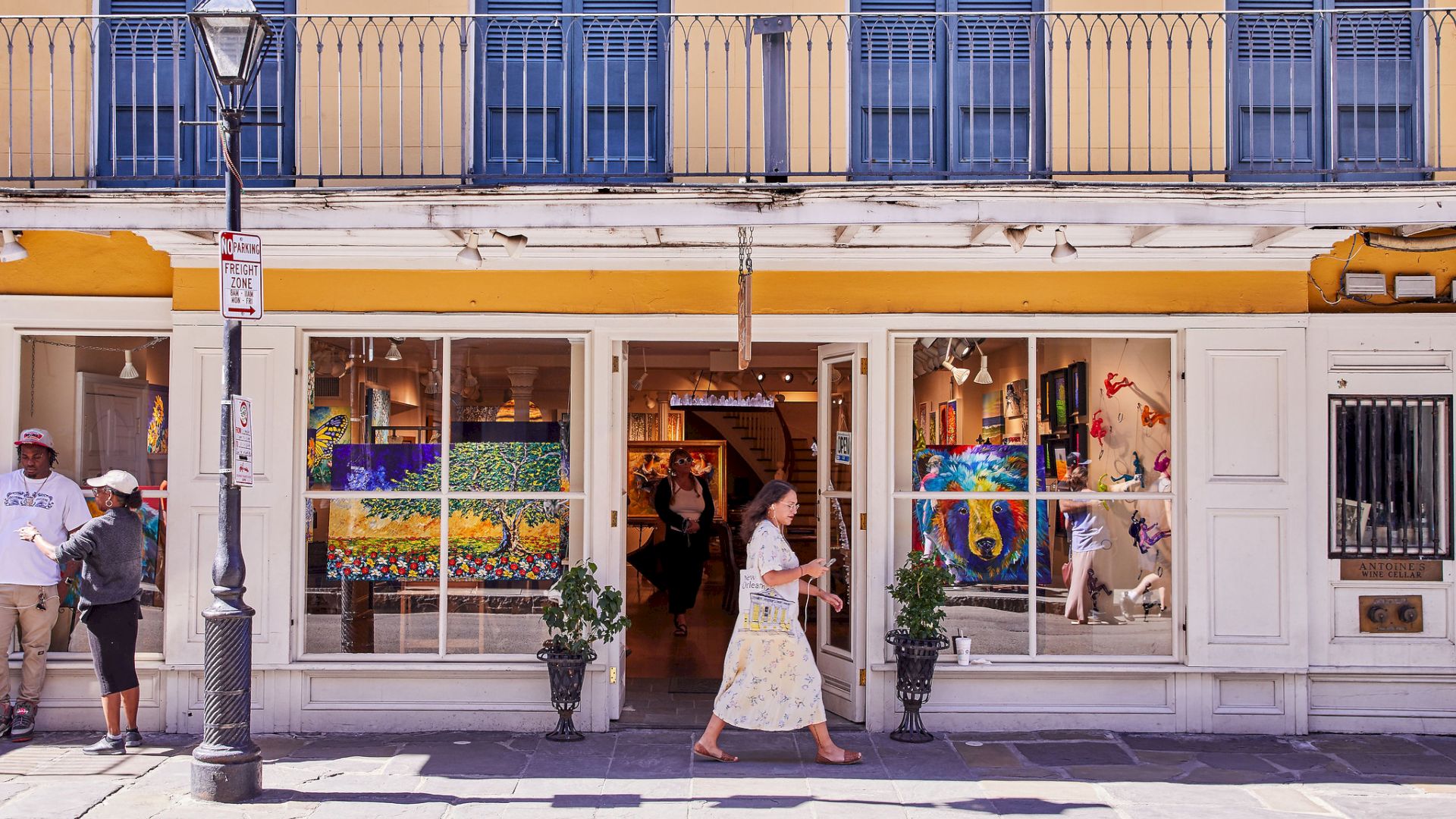 Street view of an art shop/gallery storefront with colorful paintings in the window and people walking outside.