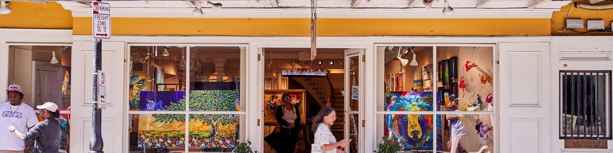 Street view of an art shop/gallery storefront with colorful paintings in the window and people walking outside.