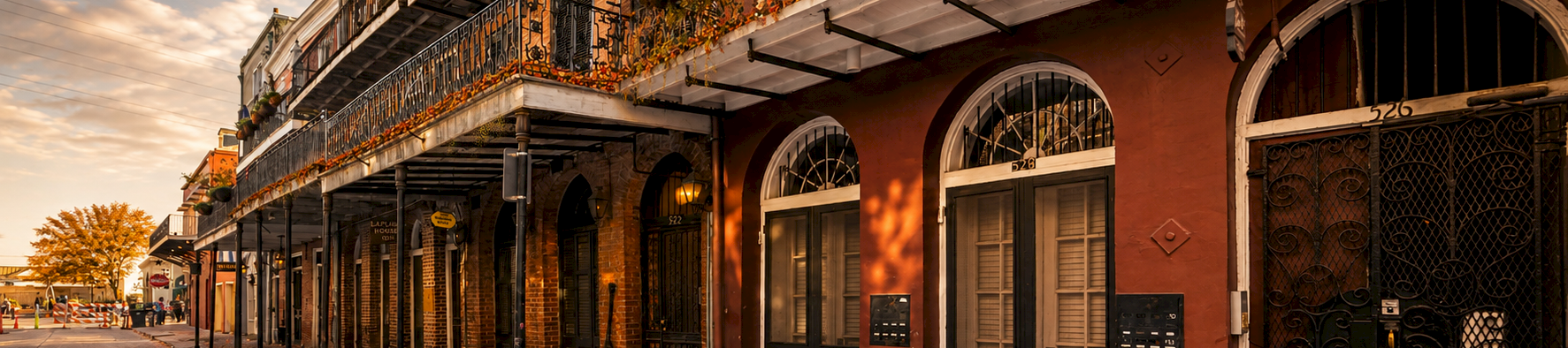 A brick courtyard building with wrought-iron balconies and arched doors, decorated with autumn plants and pumpkins along a sidewalk.