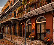 A brick courtyard building with wrought-iron balconies and arched doors, decorated with autumn plants and pumpkins along a sidewalk.
