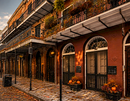 A brick courtyard building with wrought-iron balconies and arched doors, decorated with autumn plants and pumpkins along a sidewalk.