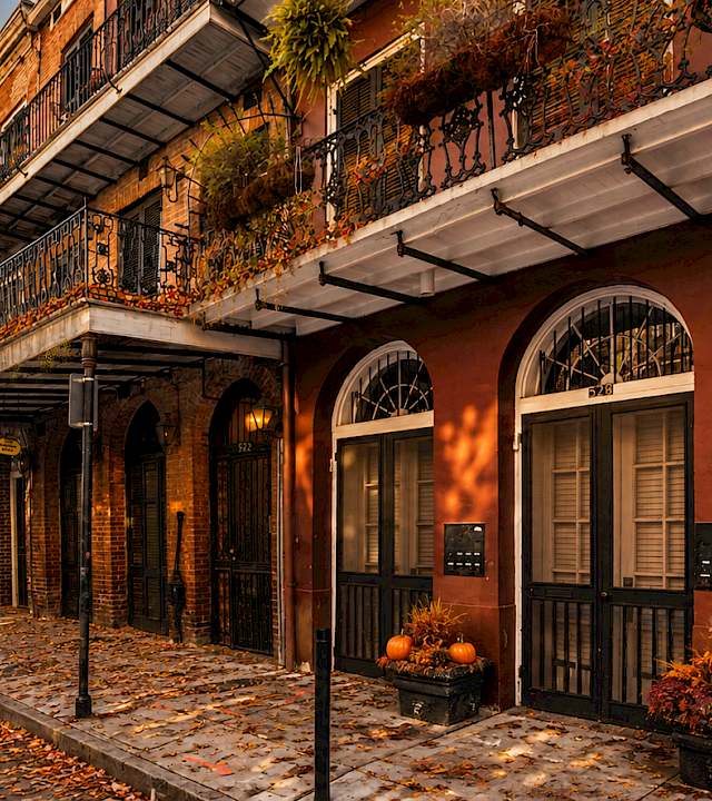 A brick courtyard building with wrought-iron balconies and arched doors, decorated with autumn plants and pumpkins along a sidewalk.
